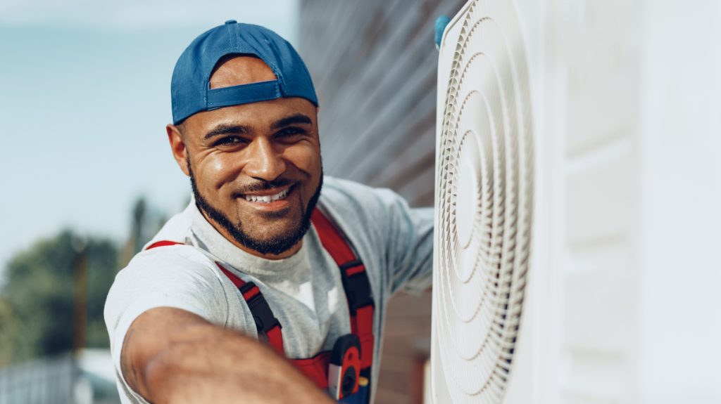 Smiling HVAC technician in a blue cap and red overalls working on an outdoor air conditioner unit on a sunny day.