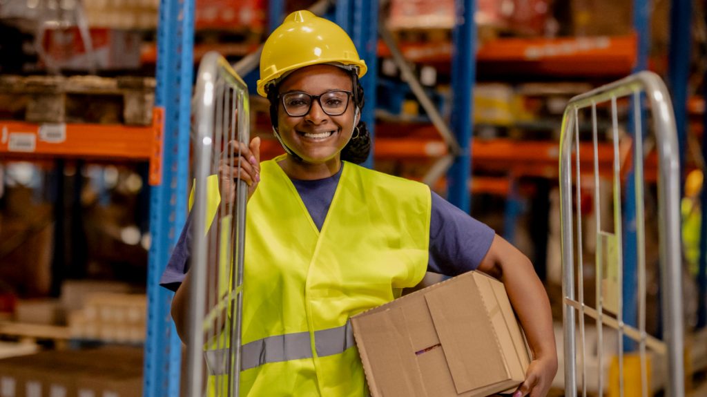 Smiling woman in a yellow hard hat and high-visibility vest stands by a cart, holding a cardboard box in a warehouse.