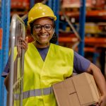 Smiling woman in a yellow hard hat and high-visibility vest stands by a cart, holding a cardboard box in a warehouse.